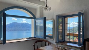 A room with a wooden table and a chair, a window with blue shutters, and a view of a lake and mountains. - Casa del Prete (Cannero Riviera)