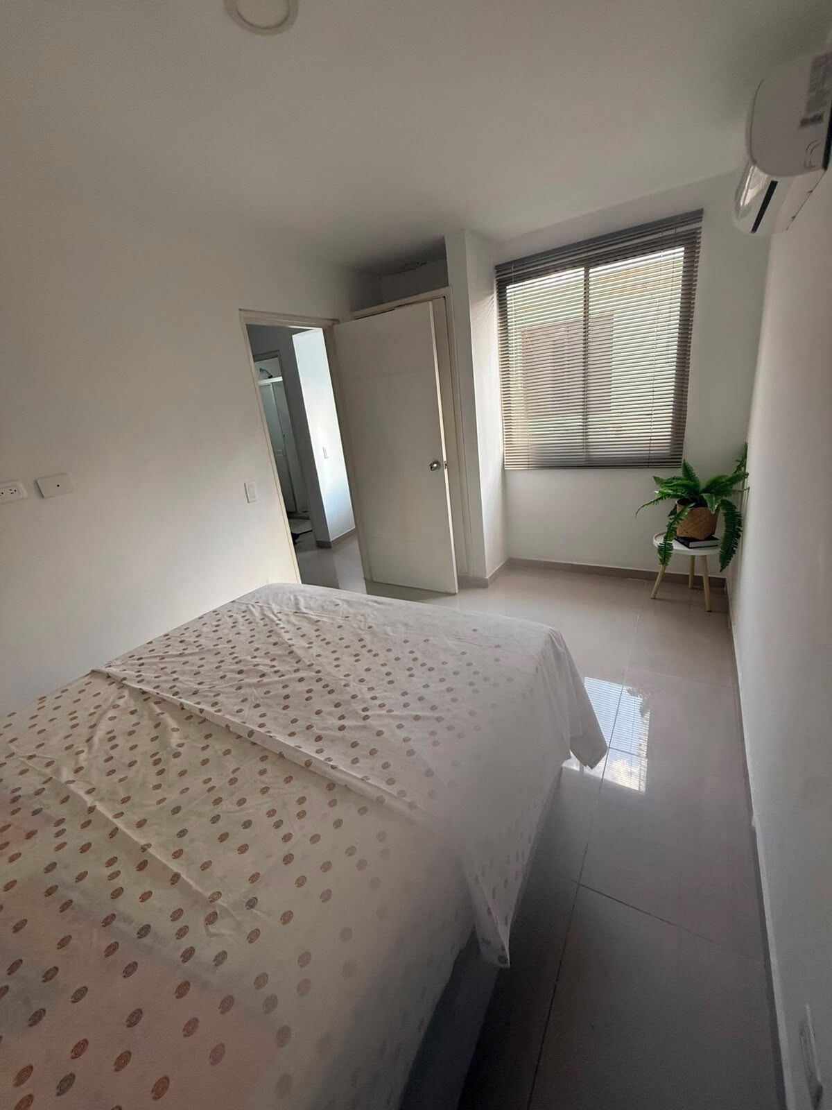 A bedroom with a bed, a wardrobe, a window with blinds, and a potted plant.