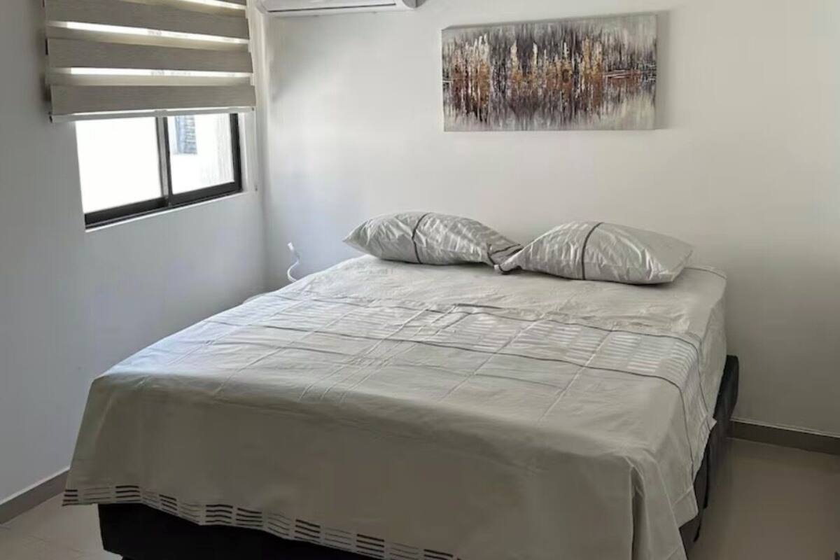 A bedroom with a bed, a window with blinds, an air conditioning unit, and a painting on the wall.