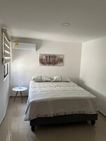 Quarto com cama, mesa de cabeceira com abajur, ar condicionado na parede e um quadro.