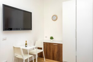 Pieni keittonurkkaus, jossa on litteänäyttöinen televisio, puinen kaappi, pieni kasvi ja seinäkello.