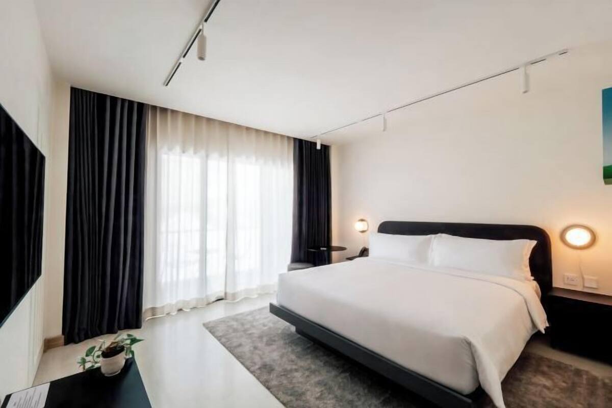 A modern hotel room with a large bed, a desk with a chair, a potted plant, and a view of the city.