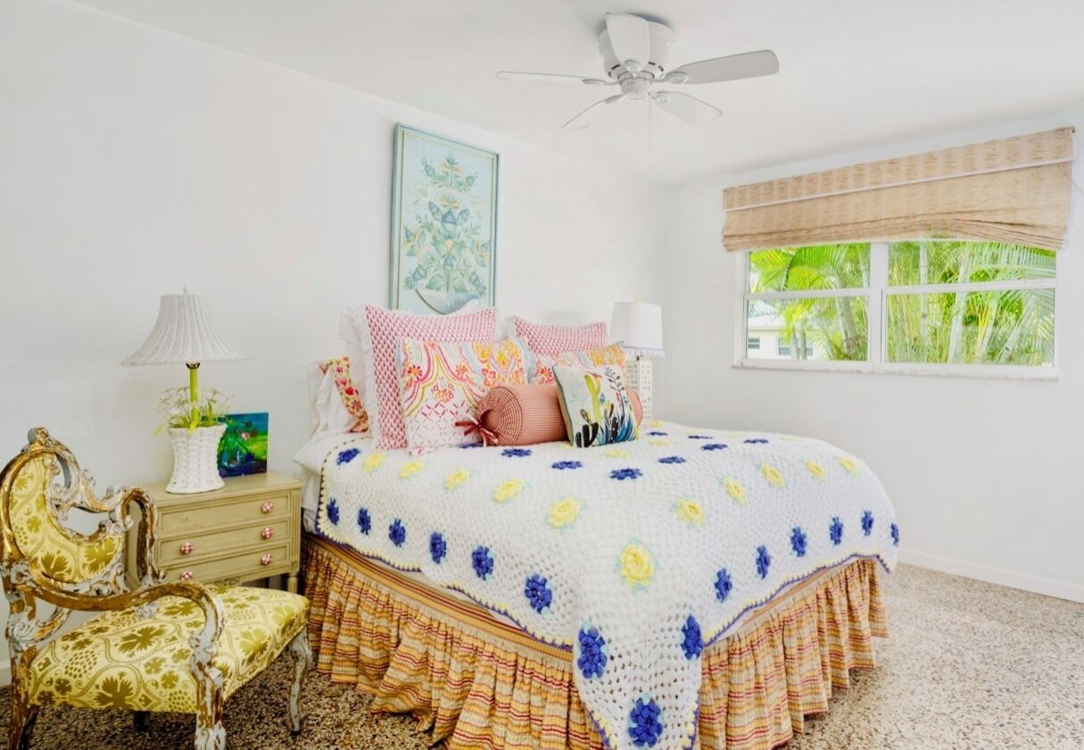 A bedroom with a bed, a chair, a nightstand, a lamp, a ceiling fan, a window with curtains, and a painting on the wall.