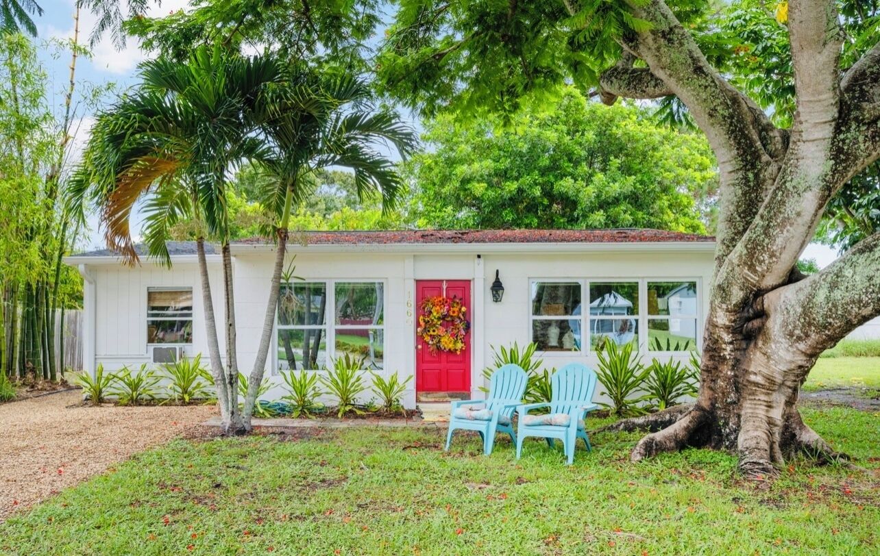 Un bâtiment blanc avec une porte rouge, entouré de plantes et d’arbres tropicaux.