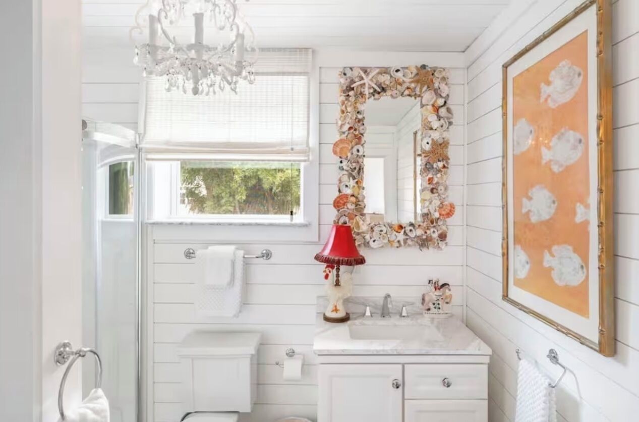 A bathroom with a white vanity, a red lamp, a decorative mirror, and a framed artwork on the wall.