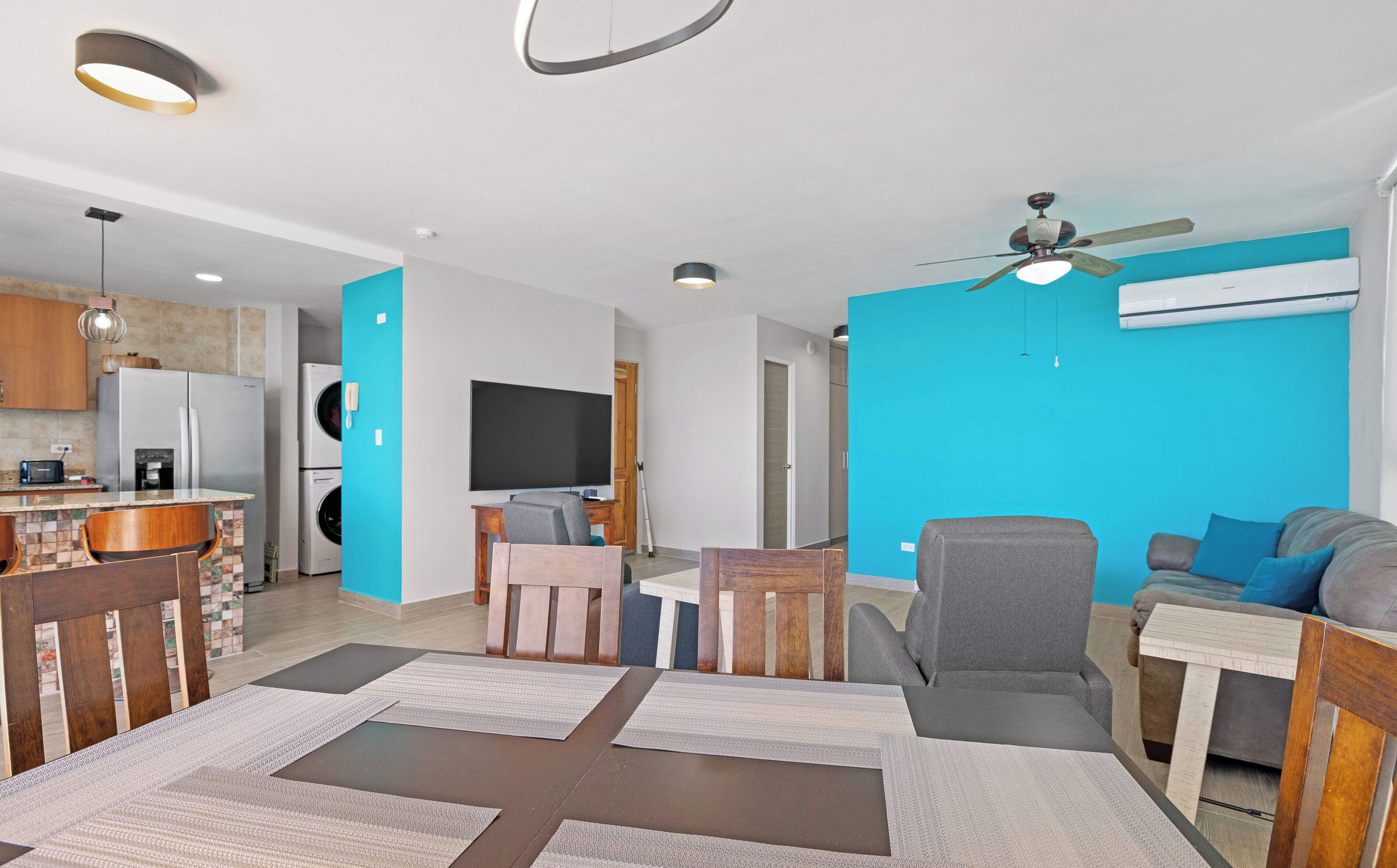 Ein modernes Wohnzimmer mit großem Fernseher, grauem Sofa und Essbereich mit Holztisch und Stühlen.