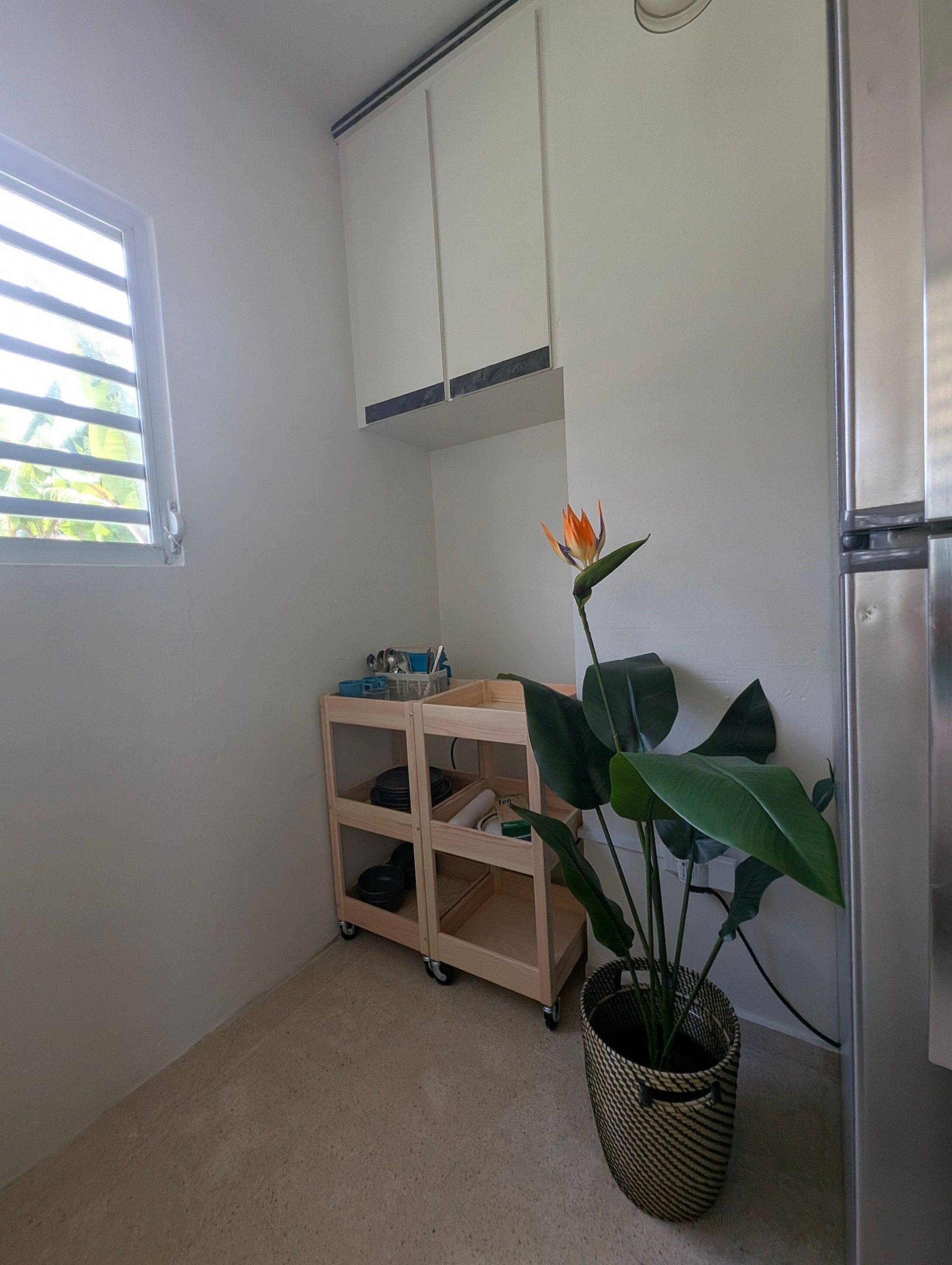 A small kitchen with white cabinets, a wooden cart with kitchenware, a potted plant, and a refrigerator.