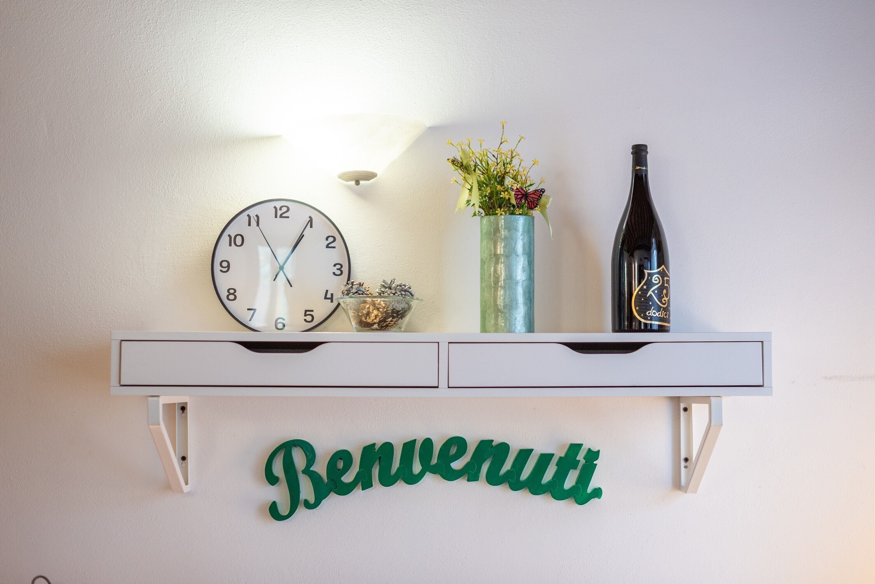 A wall clock, a bottle, a vase with flowers, and a welcome sign on a shelf.