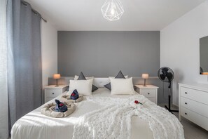 Een moderne slaapkamer met een groot bed, twee nachtkastjes met lampen, een staande ventilator en een geometrische plafondlamp.