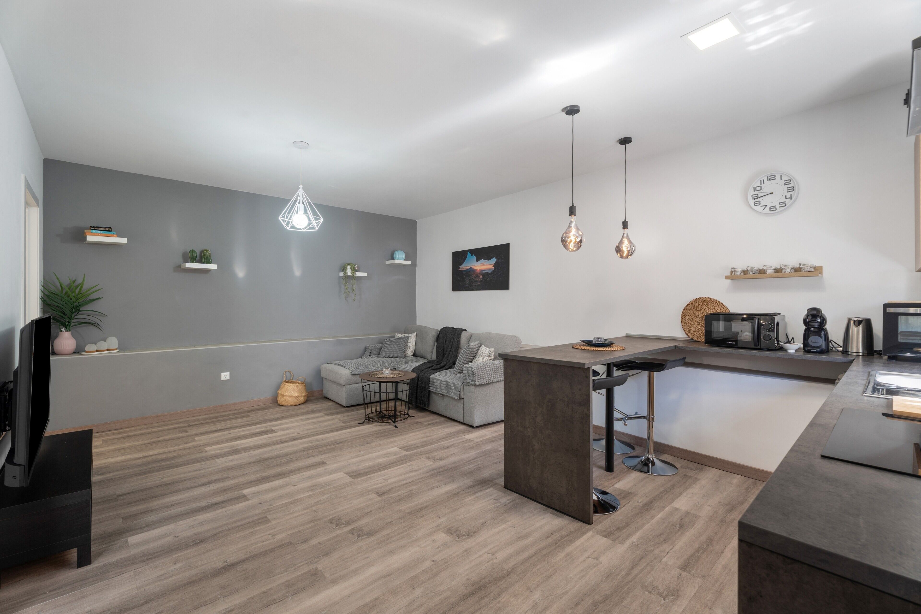 Un espacio habitable moderno con un sofá gris, una zona de comedor, una cocina con electrodomésticos y una lámpara colgante.