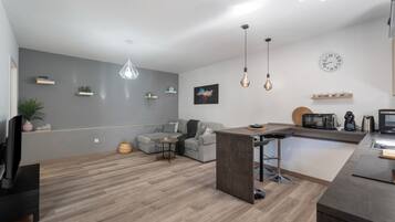 Un espacio habitable moderno con un sofá gris, una zona de comedor, una cocina con electrodomésticos y una lámpara colgante.