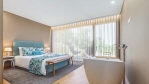 A modern bedroom with a bed, bedside tables, a bench, a large window with curtains, and a white bathtub.