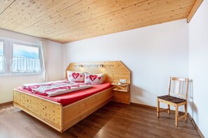 Uma cama de madeira com colchão vermelho, uma mesinha de cabeceira de madeira, uma cadeira de madeira e uma janela com cortinas.
