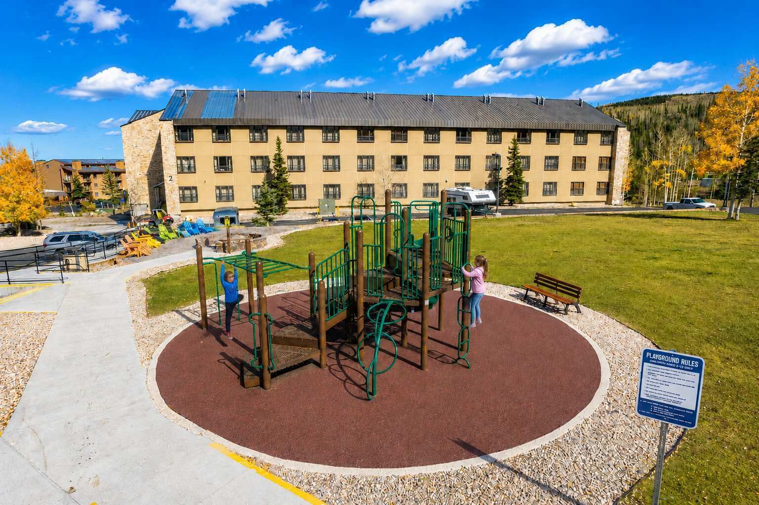 Ein Spielplatz mit Klettergerüsten und einer roten, gummierten Oberfläche, umgeben von einem befestigten Bereich und einem Gebäude im Hintergrund.
