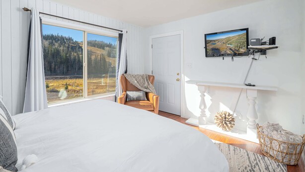 Ein Schlafzimmer mit einem großen Bett, einem Sessel am Fenster, einem Fernseher an der Wand und Blick auf einen Wald.