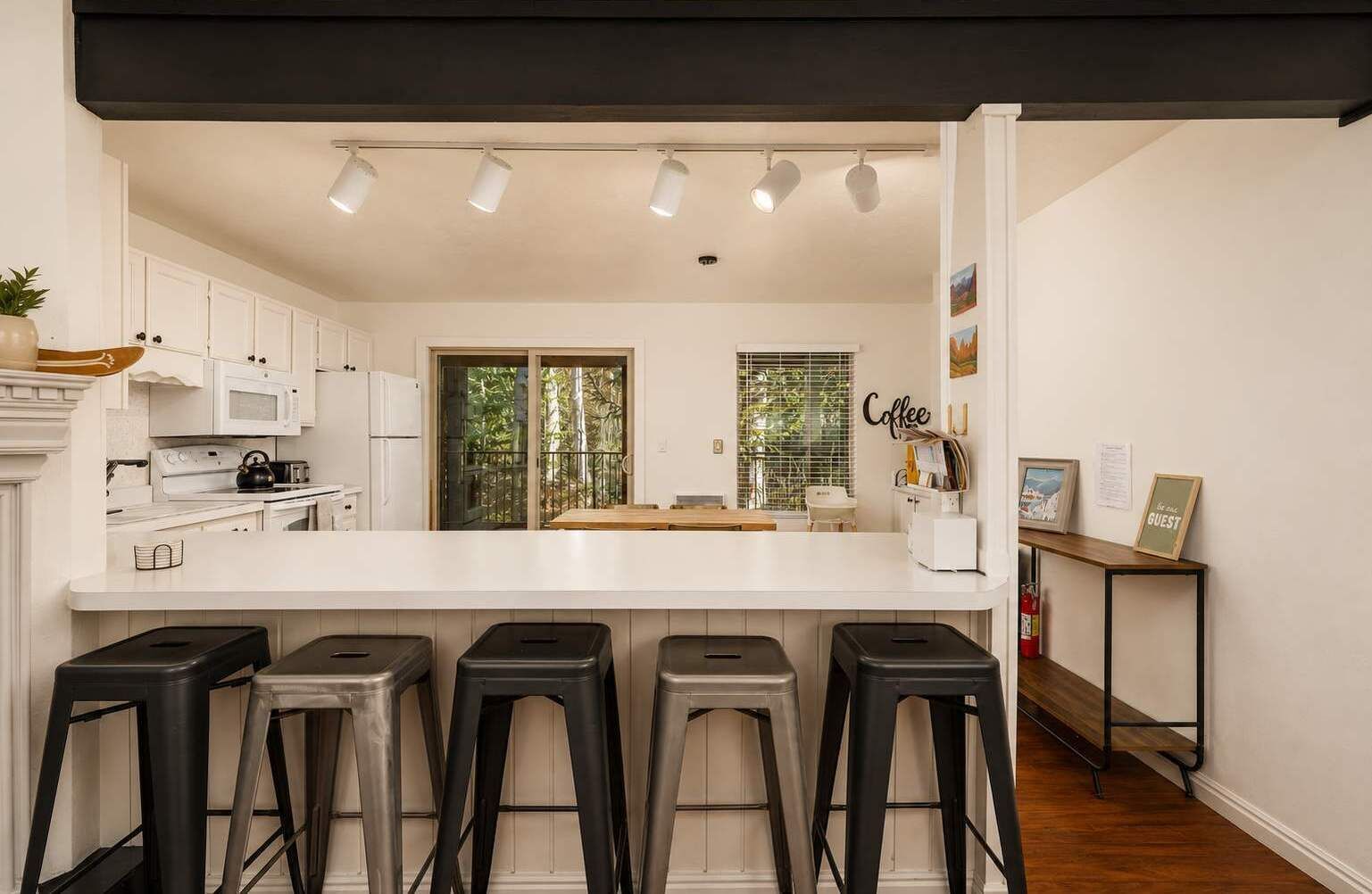 A modern kitchen with white cabinets, a central island with bar stools, and a dining area visible through a glass door.