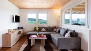 Een moderne woonkamer met een houten plafond, een grijze bank, een tafel met een vaas en fruit, en een televisie op een kast.