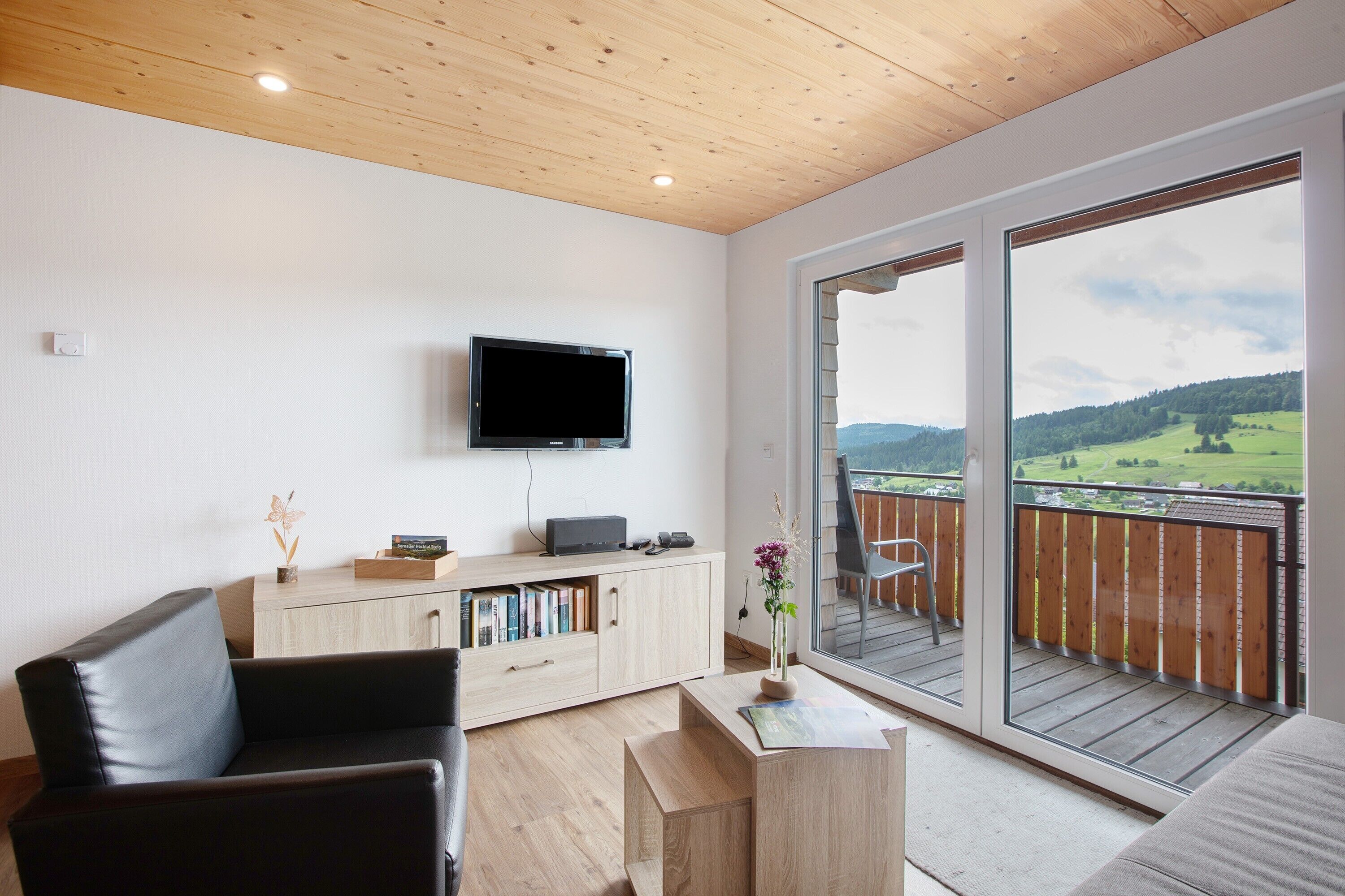 A modern living room with a flat-screen TV, a wooden ceiling, a balcony with a view of the countryside, and a cozy seating area.