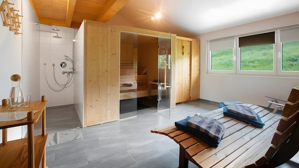 Moderni huone, jossa on puinen katto, sauna, lasinen suihkuseinä, puinen penkki ja suuret ikkunat.