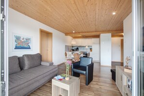 Una sala de estar moderna con un sofá, una mesa de centro y una silla. Se observa una zona de cocina al fondo con armarios y una mesa de comedor.