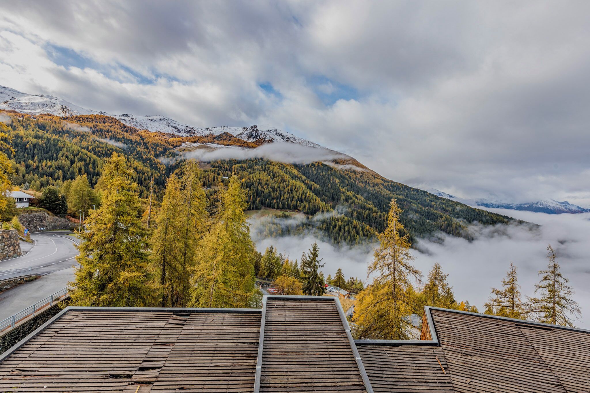 A mountain landscape with a road, trees, and a building with a sloped roof.