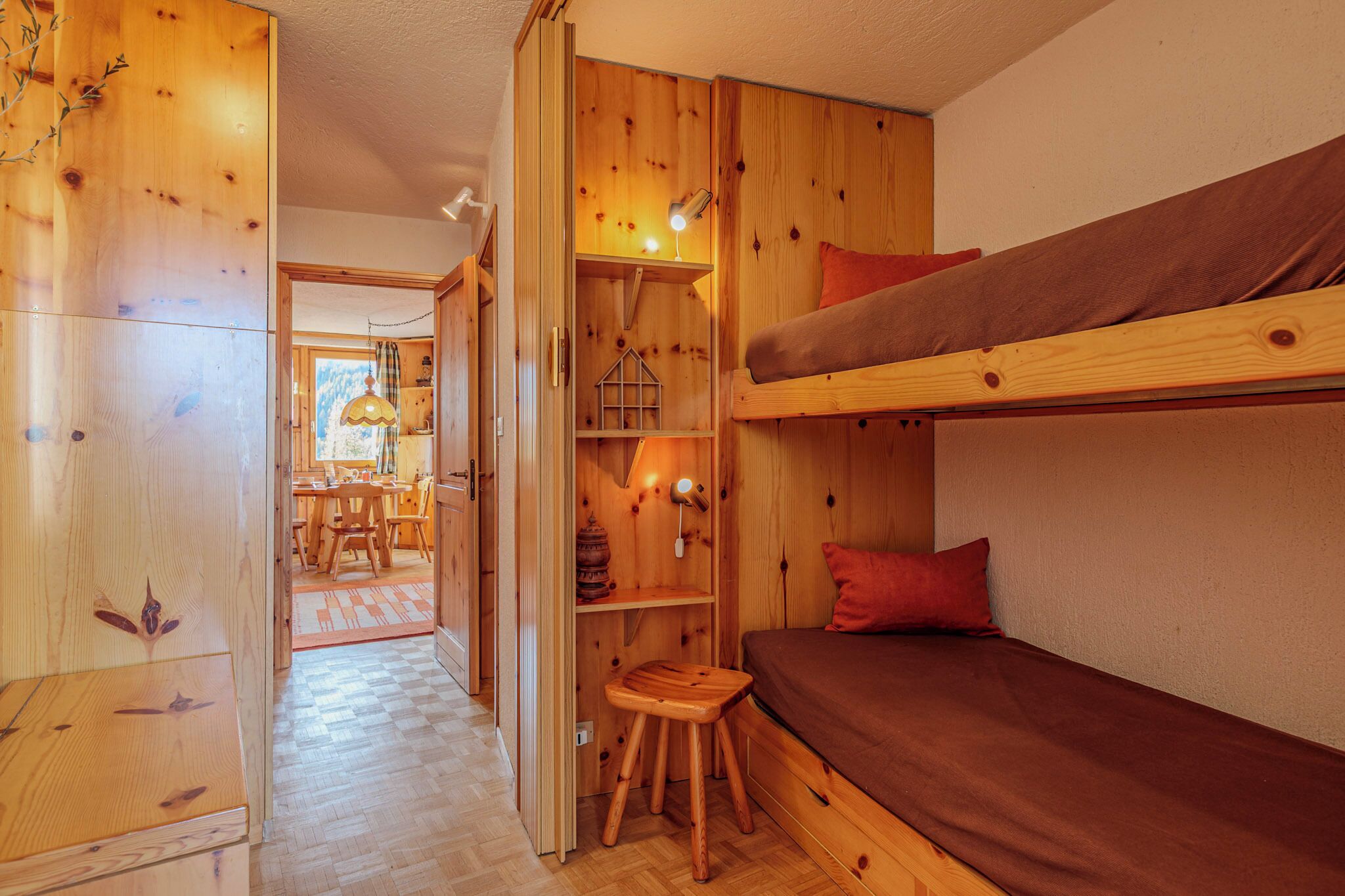 A wooden cabin interior with a bunk bed, a small wooden stool, and a dining area visible through an open door.