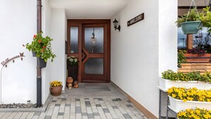 A building entrance with a brown door, a welcome sign, a potted plant, and a hanging planter with flowers. - Haus Hochtalblick Apartment "Rechberg" with Panoramic View, Wi-Fi, DreiWelten-Card Included (Bernau im Schwarzwald)