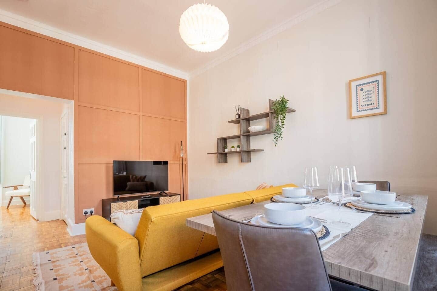 A modern living room with a yellow sofa, a dining table set with white plates and glasses, a TV, and wooden cabinets.