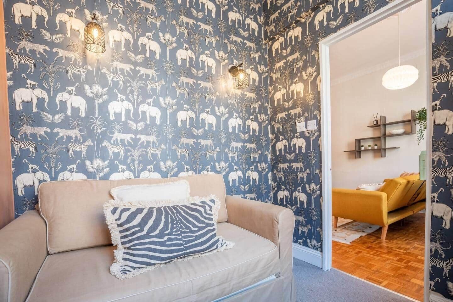 A room with a patterned wallpaper featuring animal prints, a beige sofa with a zebra-patterned pillow, and a yellow armchair.