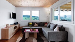 A modern living room with a wooden ceiling, a grey sofa, a wooden cabinet, a television, and a large window with a view of green fields. - Hochtalblick: Apartment "Blösling 1" with Panoramic View, Wi-Fi & DreiWelten-Card Included (Bernau im Schwarzwald)