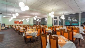 A spacious dining area with multiple tables and chairs, arranged in rows, with overhead lighting fixtures.