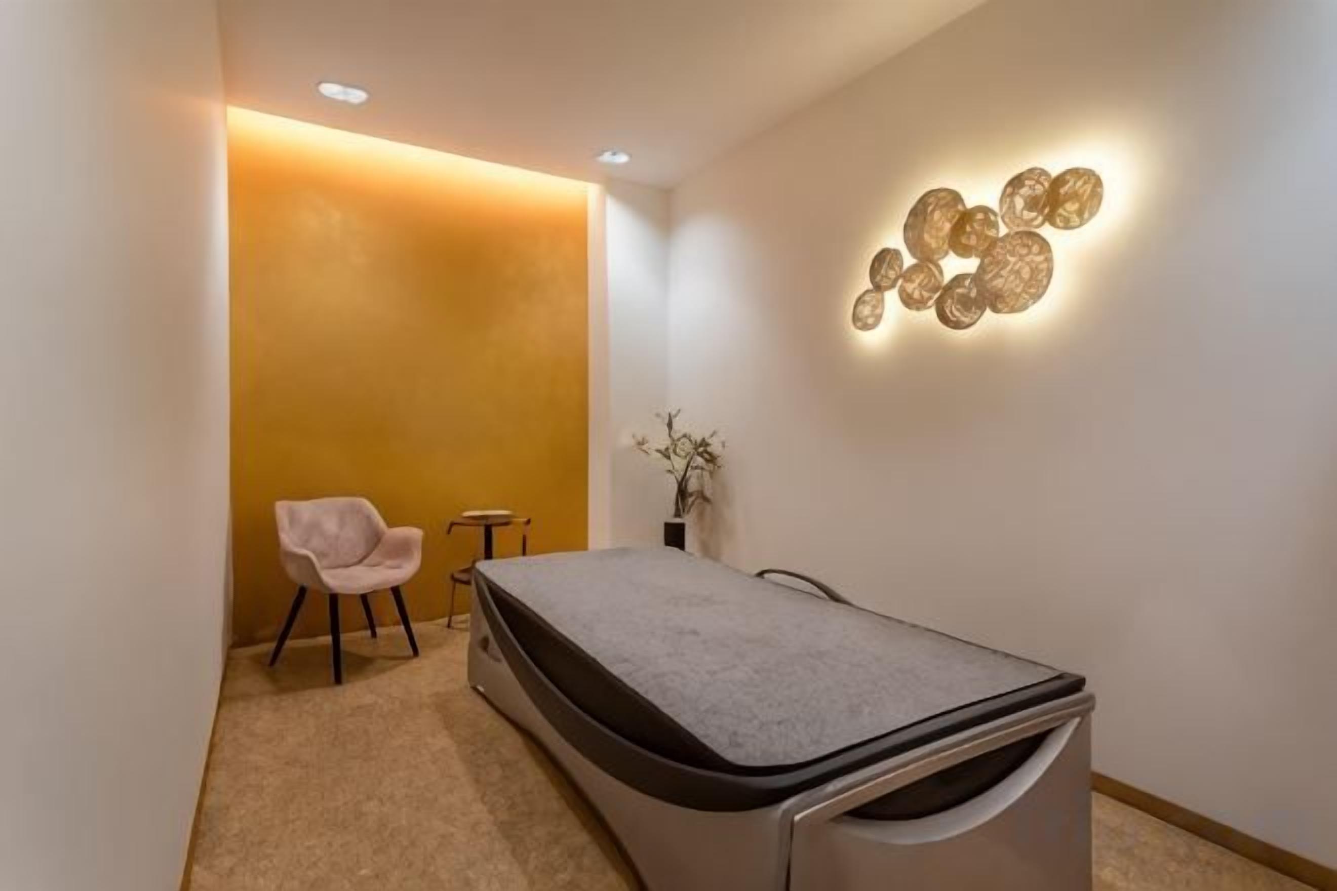 Une pièce comprenant une table de massage, une chaise, une petite table et un luminaire fixé au mur.