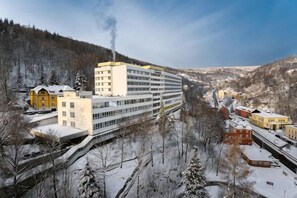 A multi-story building with a central atrium, surrounded by snow-covered trees and a mountainous backdrop. - Hotel Behounek (Karlovy Vary District)