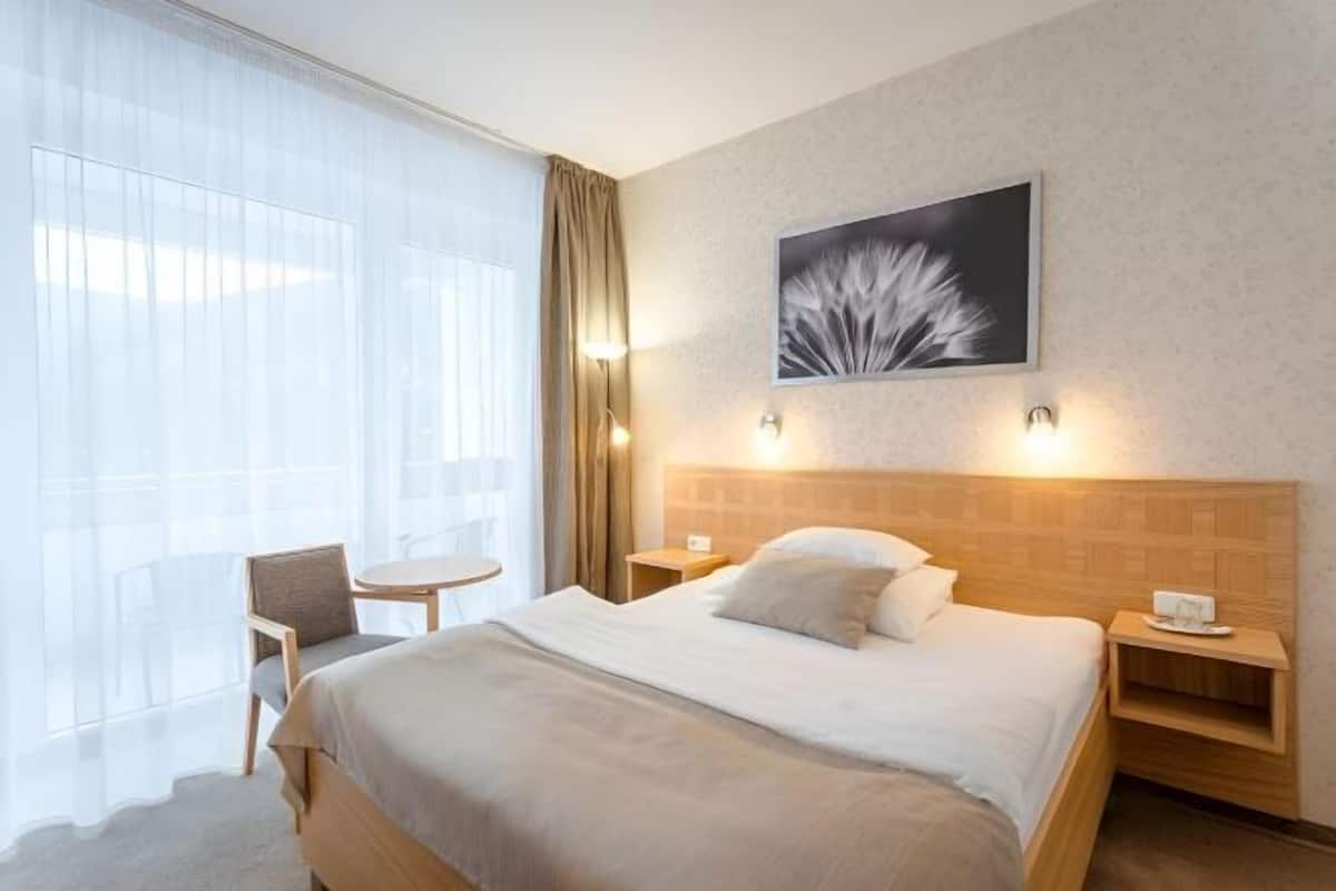 A hotel room with a large bed, a chair, a small table, and a wall-mounted picture.
