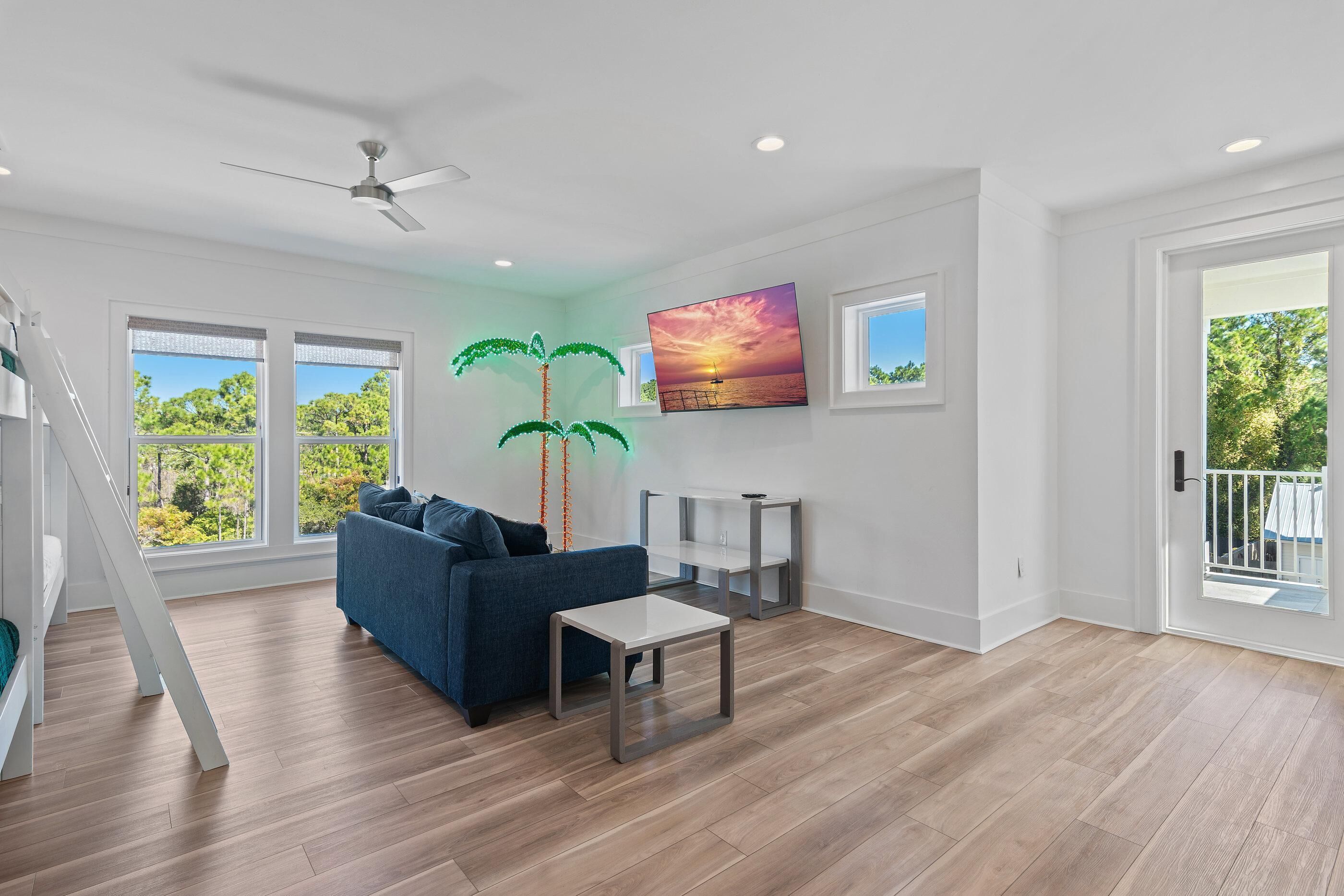 A spacious living room with a blue sofa, a small table, a ceiling fan, and a large window with a view of trees.