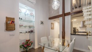 Een eethoek met een glazen tafel en stoelen, een glazen plank met decoratieve objecten, een geweven mand met boeken en een vaas met bloemen.