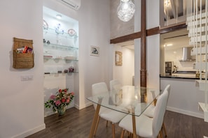 Un comedor con una mesa y sillas de vidrio, un estante de vidrio con objetos decorativos, una cesta tejida con libros y un jarrón con flores.