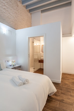 Un dormitorio con cama, mesita de noche, lámpara, baño con ducha y suelo de madera.
