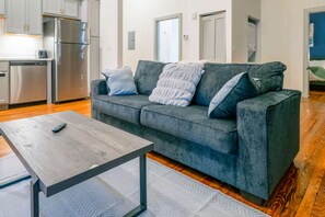 Een moderne woonkamer met een grijze bank, een houten salontafel, en een keuken met roestvrijstalen apparatuur.