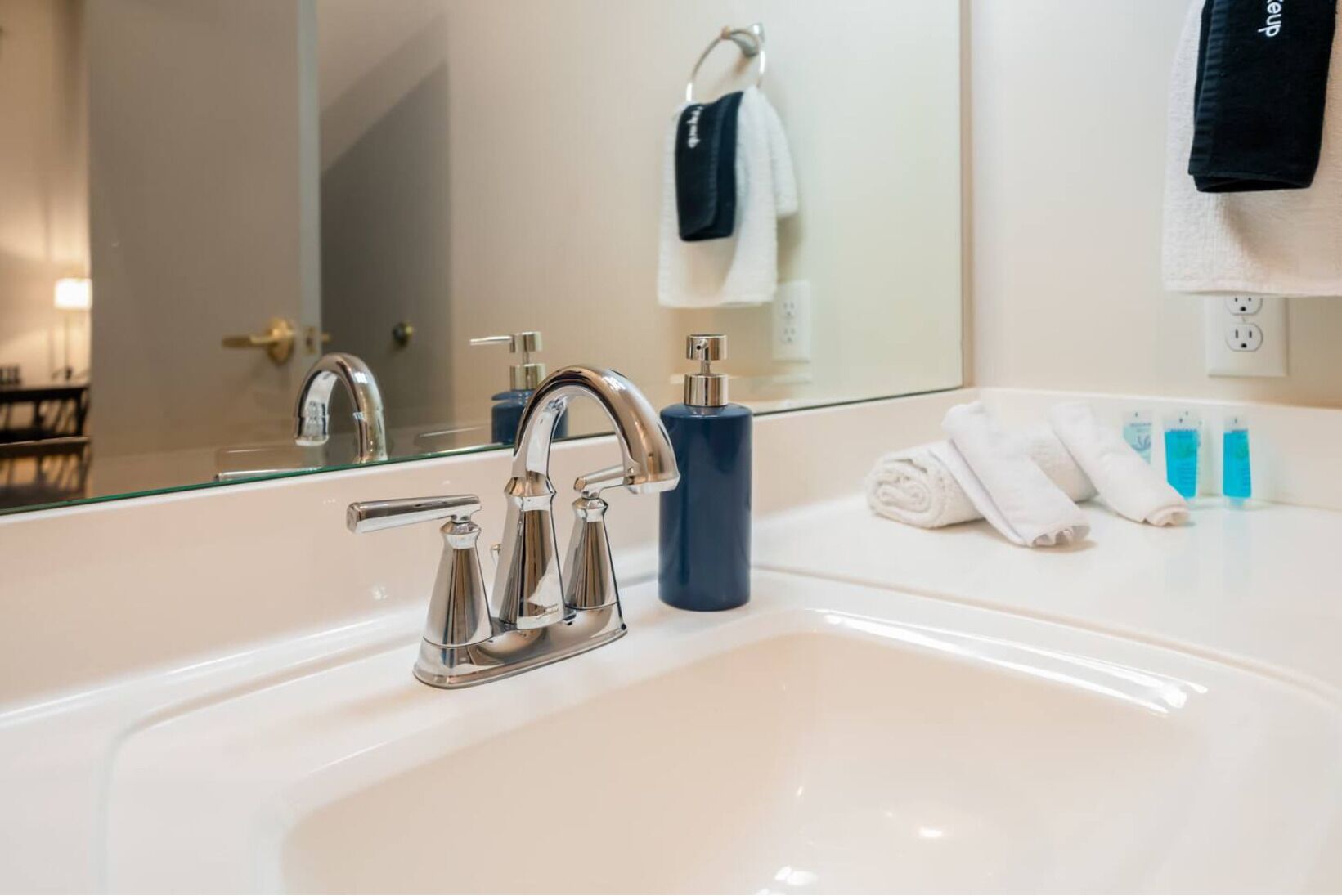 A modern bathroom with a white sink, chrome faucets, and a large mirror. There are towels and toiletries on the counter.