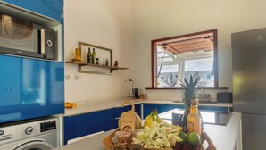 Eine kompakte Küche mit Waschmaschine, Mikrowelle und Kühlschrank. Ein Holzregal mit Flaschen und ein Fenster über der Spüle.