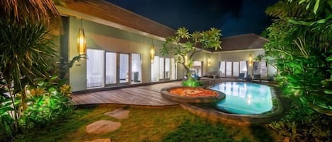 A well-lit outdoor area with a swimming pool, wooden deck, and surrounding greenery.