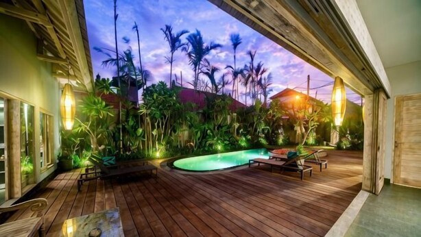 Una terraza de madera con piscina, rodeada de plantas tropicales y vista al atardecer.
