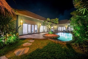 Une vue nocturne d’un complexe hôtelier avec une piscine, une terrasse en bois et une végétation luxuriante.