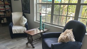 Una habitación con un ventanal grande, un sillón blanco, otro sillón oscuro, una mesa de centro de madera y una estantería llena de libros.