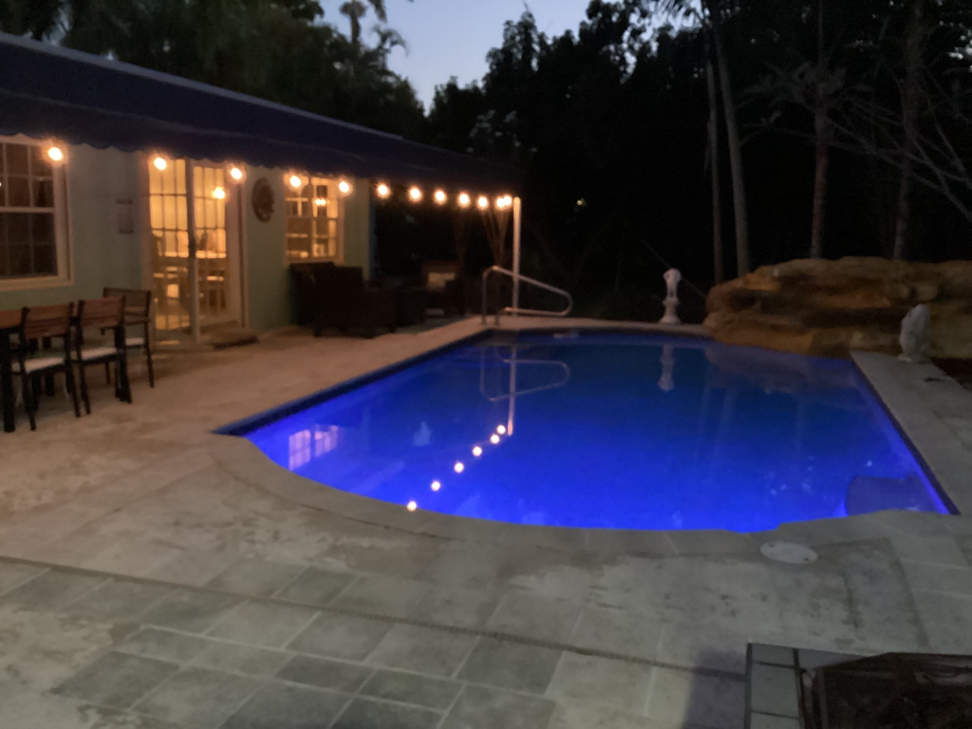 A swimming pool with a blue tint, surrounded by a patio area with chairs and a table.