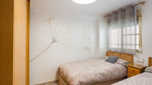 Slaapkamer met twee bedden, een houten hoofdbord, een nachtkastje, een wand met bloemrijk decor en een raam met jaloezieën.