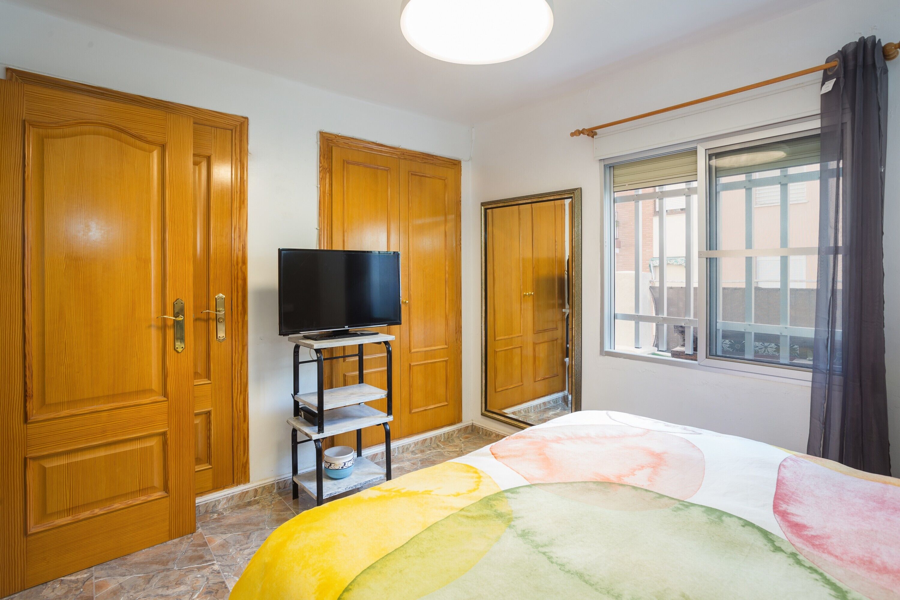 A bedroom with wooden wardrobes, a television on a stand, a bed with a colorful blanket, and a window with curtains.