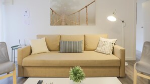Ein Wohnzimmer mit einem beigefarbenen Sofa, einem kleinen Tisch mit einer Pflanze, einem hängenden Kunstwerk, das eine Brücke zeigt, und einer Stehlampe.