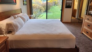 Une chambre avec un grand lit, une tête de lit en bois, une table de chevet, une commode et une porte-fenêtre coulissante donnant sur un jardin.